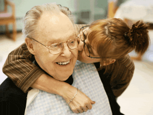 Elderly man hugged and kissed affectionately by woman.